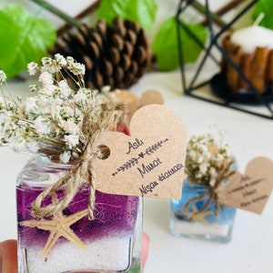 Puede incluir: Un peque&ntilde;o frasco de vidrio lleno de arena morada y blanca y una estrella de mar. El frasco est&aacute; atado con un cordel y una peque&ntilde;a flor blanca. Una etiqueta en forma de coraz&oacute;n est&aacute; unida al frasco con el texto "Aslı & Murat Nişan Hatırası".