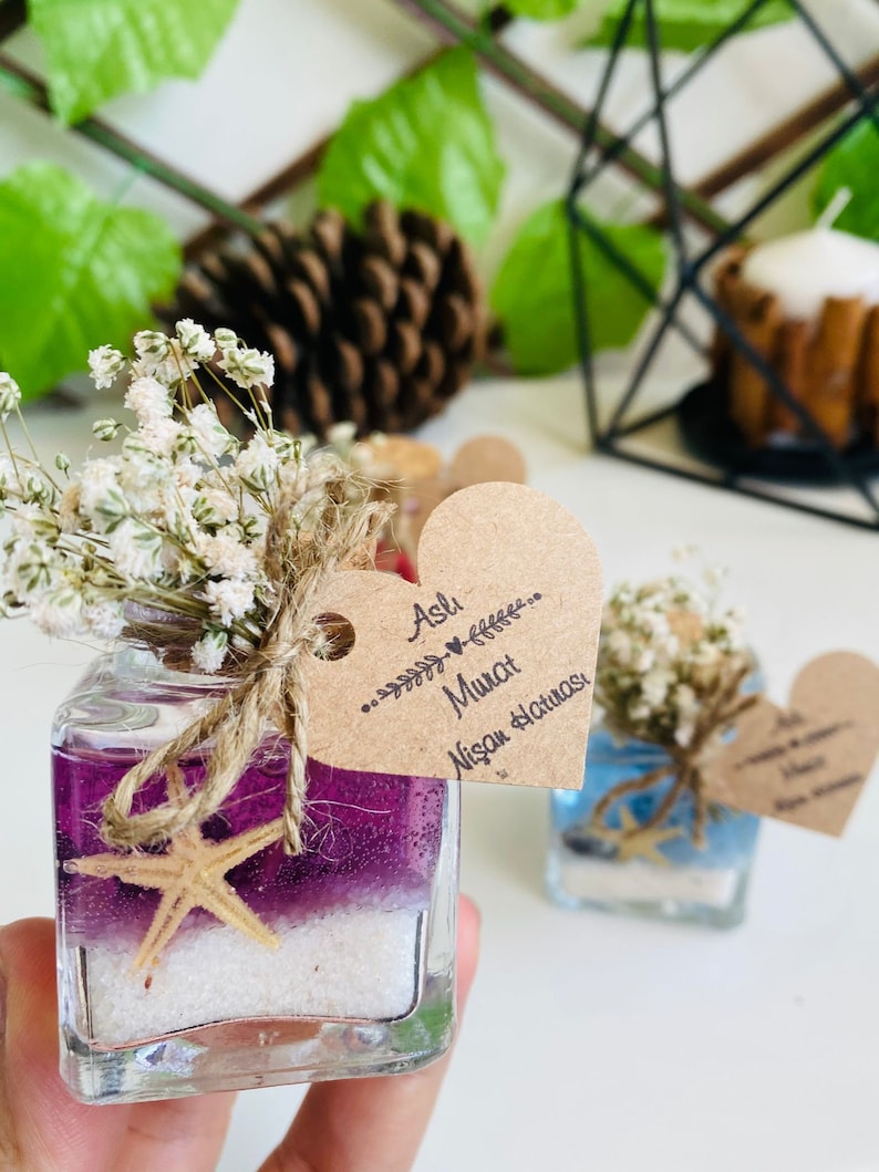 Puede incluir: Un peque&ntilde;o frasco de vidrio lleno de arena morada y blanca y una estrella de mar. El frasco est&aacute; atado con un cordel y una peque&ntilde;a flor blanca. Una etiqueta en forma de coraz&oacute;n est&aacute; unida al frasco con el texto "Aslı & Murat Nişan Hatırası".