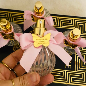 Pode incluir: Conjunto de quatro frascos de perfume de vidro transparente com bicos de pulverização dourados e laços cor-de-rosa. Cada frasco tem um amuleto de borboleta dourado com o texto "Batizado da Tiara 30.07.2023".