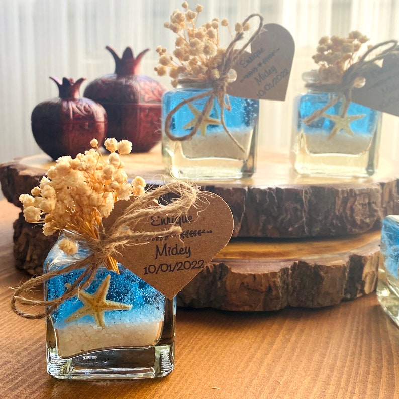 Puede incluir: Peque&ntilde;os frascos de vidrio llenos de arena azul y una estrella de mar, decorados con flores blancas secas y una etiqueta en forma de coraz&oacute;n marr&oacute;n con el texto "Enrique & Midey 10/01/2022".