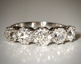 May include: A silver band ring with five round, clear diamonds set in a row. The diamonds are surrounded by smaller diamonds.