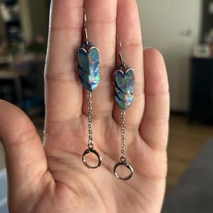 Op de afbeelding: Hangende oorbellen met iriserende blauw-groene ovale hangers. De hangers zijn verbonden met zilverkleurige kettingen die eindigen in kleine ringen. Deze oorbellen zijn ontworpen voor doorboorde oren.