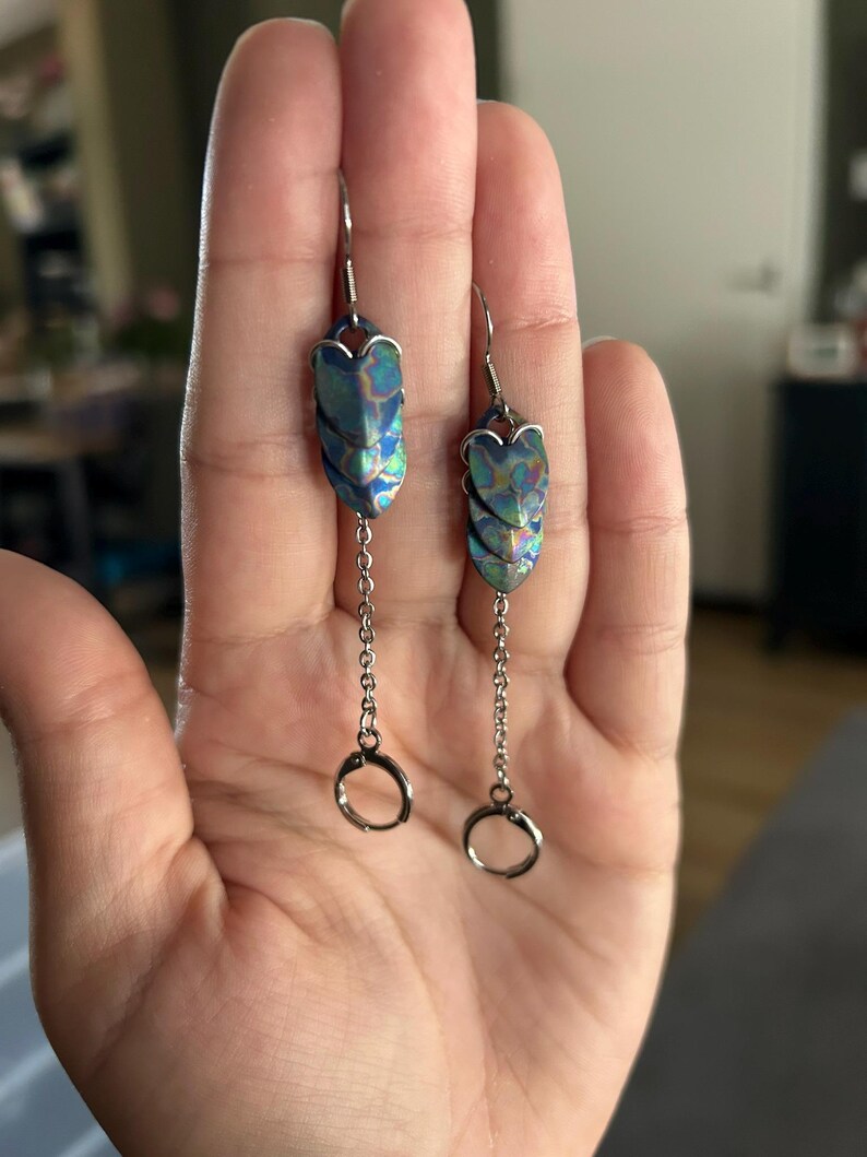 Op de afbeelding: Hangende oorbellen met iriserende blauw-groene ovale hangers. De hangers zijn verbonden met zilverkleurige kettingen die eindigen in kleine ringen. Deze oorbellen zijn ontworpen voor doorboorde oren.