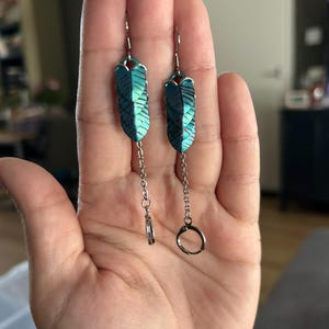 Op de afbeelding: Een paar turquoise en zwarte oorringen in de vorm van een veer. De oorbellen hebben een zilverkleurige haak en ketting, met een kleine ring aan de onderkant van &eacute;&eacute;n oorbel en een grotere ring aan de andere. De oorbellen worden in een hand gehouden.