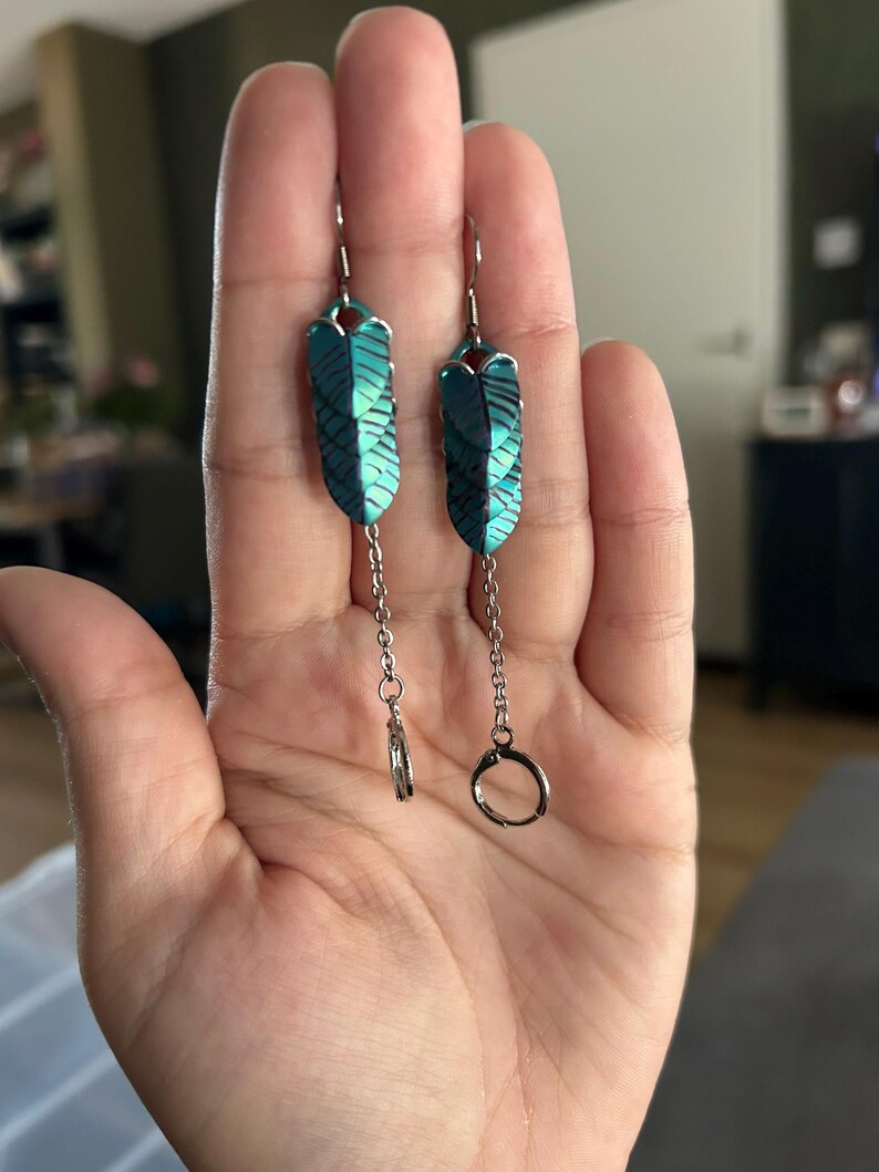 Op de afbeelding: Een paar turquoise en zwarte oorringen in de vorm van een veer. De oorbellen hebben een zilverkleurige haak en ketting, met een kleine ring aan de onderkant van &eacute;&eacute;n oorbel en een grotere ring aan de andere. De oorbellen worden in een hand gehouden.