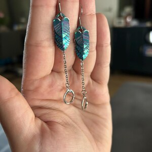 Op de afbeelding: Een paar oorhangers met een veervormig ontwerp. De oorbellen hebben een iriserende kleurstelling in teal en paars. Elke oorbel heeft een kleine ketting met een zilverkleurige ring aan het uiteinde. De oorbellen worden in een hand gehouden.
