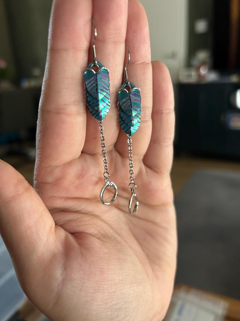 Op de afbeelding: Een paar oorhangers met een veervormig ontwerp. De oorbellen hebben een iriserende kleurstelling in teal en paars. Elke oorbel heeft een kleine ketting met een zilverkleurige ring aan het uiteinde. De oorbellen worden in een hand gehouden.