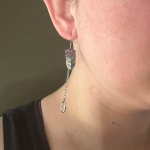 Op de afbeelding: Een bungelende oorbel met een zilveren ketting en een decoratief, gelaagd, bladachtig ontwerp in roze, groen en paars. De oorbel hangt aan een zilveren haak en heeft onderaan een kleine, zilveren ring.