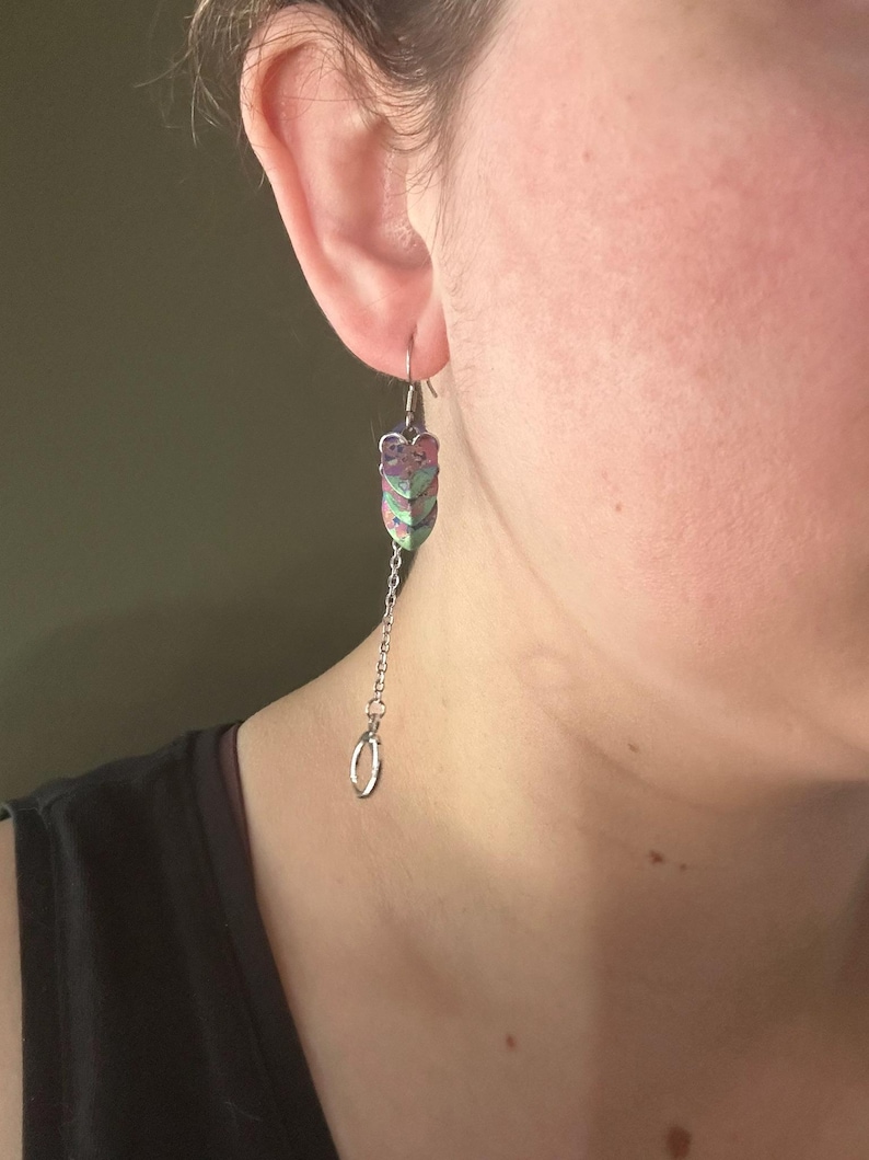 Op de afbeelding: Een bungelende oorbel met een zilveren ketting en een decoratief, gelaagd, bladachtig ontwerp in roze, groen en paars. De oorbel hangt aan een zilveren haak en heeft onderaan een kleine, zilveren ring.