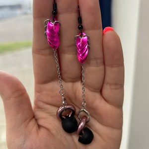 Op de afbeelding: Een paar roze hartvormige oorbellen met een ketting en zwarte oordop-achtige bedels.