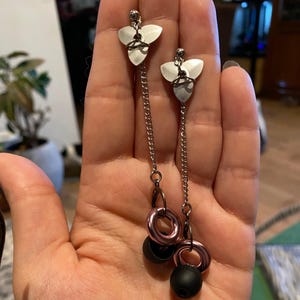 May include: A pair of silver-toned earrings with a unique design. Each earring features a three-petal top, a chain, a pink ring, and a black sphere. The earrings are displayed against a neutral background.