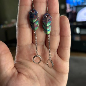 Op de afbeelding: Een paar oorhangers met een uniek ontwerp. Elke oorbel heeft iriserende, schubachtige vormen in blauw, groen en paars. Ze hangen aan zilverkleurige haken en kettingen, met een kleine ring aan de onderkant.