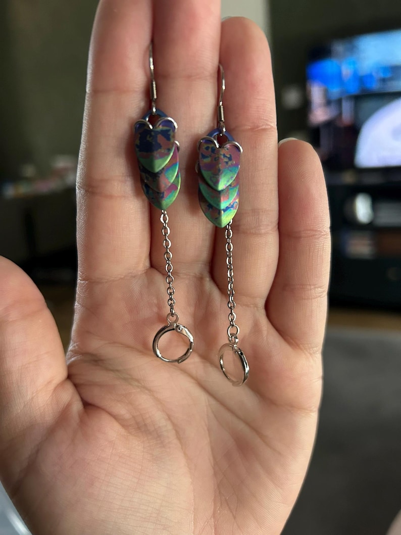 Op de afbeelding: Een paar oorhangers met een uniek ontwerp. Elke oorbel heeft iriserende, schubachtige vormen in blauw, groen en paars. Ze hangen aan zilverkleurige haken en kettingen, met een kleine ring aan de onderkant.