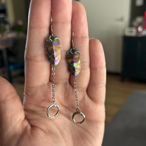 Op de afbeelding: Een paar iriserende oorbellen met een gelaagd, schubachtig ontwerp in tinten groen, paars en goud. Elke oorbel hangt aan een zilveren ketting die eindigt in een kleine, ronde ring.