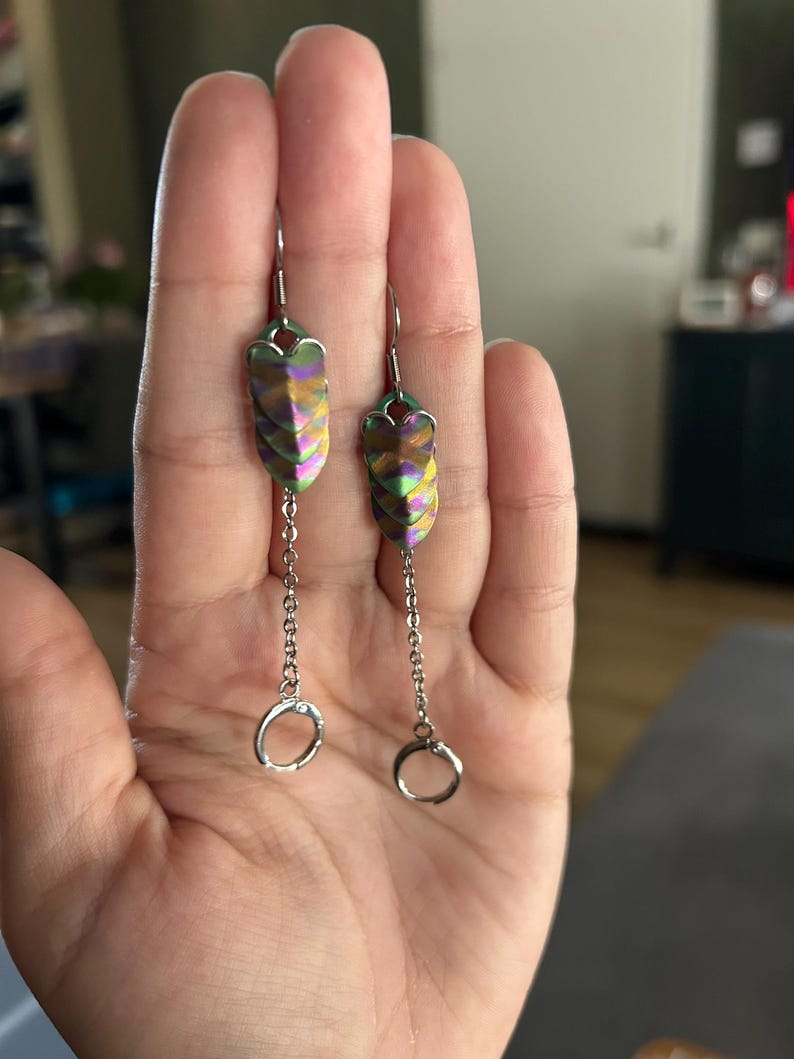 Op de afbeelding: Een paar iriserende oorbellen met een gelaagd, schubachtig ontwerp in tinten groen, paars en goud. Elke oorbel hangt aan een zilveren ketting die eindigt in een kleine, ronde ring.