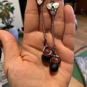 Op de afbeelding: Hangende oorbellen met een zilverkleurig metalen ontwerp. Elke oorbel heeft een bloemvormige bovenkant, een ketting en een cluster van roségouden ringen en zwarte kralen. De oorbellen worden gepresenteerd tegen een neutrale achtergrond.
