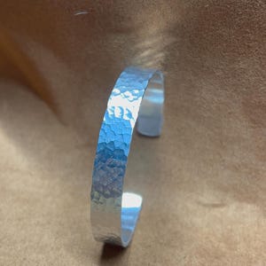 Può includere: Un bracciale a polsino argentato con una texture martellata. Il bracciale aperto è realizzato in metallo lucido e presenta una superficie leggermente irregolare. Il bracciale è mostrato su uno sfondo marrone.