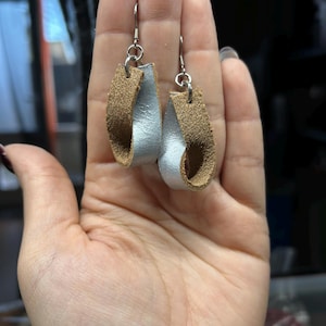 以下が含まれることがあります： ユニークなデザインの手作りイヤリング。各イヤリングは、銀色の涙の形をした要素が、質感のあるタン色の革で縁取られています。イヤリングは銀色のフックから吊り下げられています。