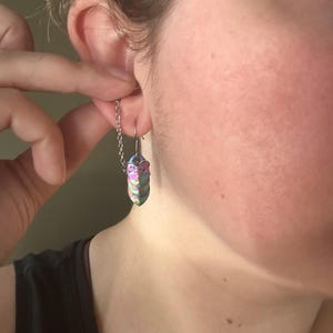 Op de afbeelding: Een close-up van een persoon die een zilveren kettingoorbel draagt met een kleurrijke, iriserende, kristalvormige hanger. De hanger heeft tinten paars, groen en blauw. De oorbel is met een haak aan het oor bevestigd.