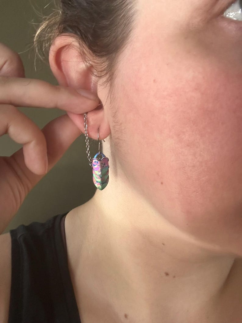 Op de afbeelding: Een close-up van een persoon die een zilveren kettingoorbel draagt met een kleurrijke, iriserende, kristalvormige hanger. De hanger heeft tinten paars, groen en blauw. De oorbel is met een haak aan het oor bevestigd.