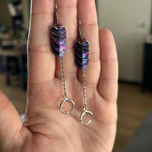 Op de afbeelding: Een paar bungelende oorbellen met een uniek ontwerp. De oorbellen hebben iriserende, schubachtige segmenten in paars, blauw en zwart. Een fijne zilveren ketting hangt een kleine, open cirkel aan de onderkant van elke oorbel.
