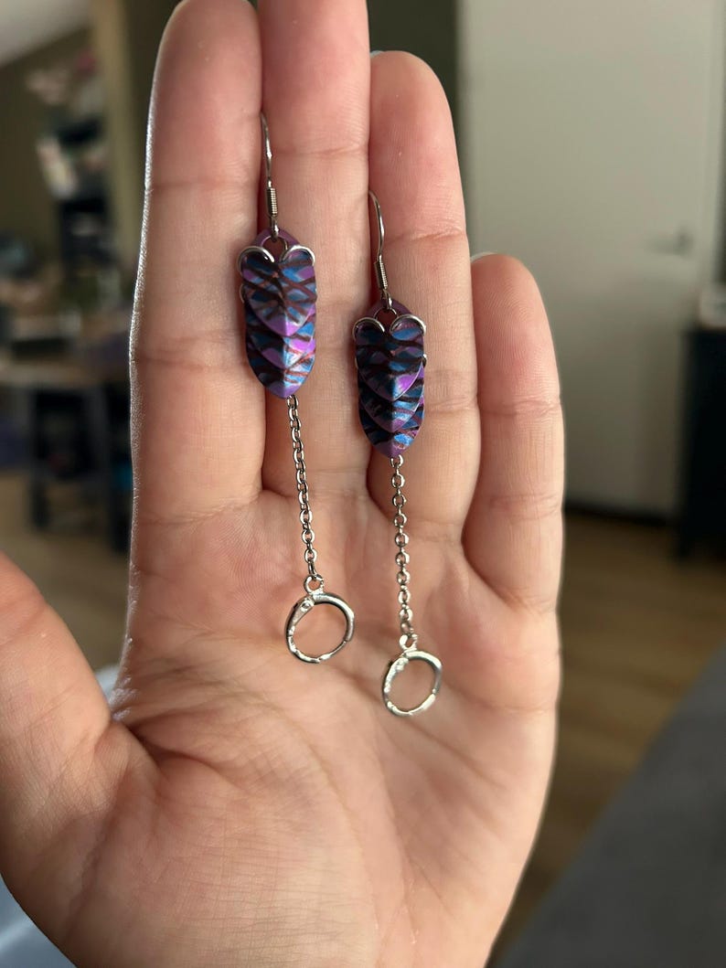 Op de afbeelding: Een paar bungelende oorbellen met een uniek ontwerp. De oorbellen hebben iriserende, schubachtige segmenten in paars, blauw en zwart. Een fijne zilveren ketting hangt een kleine, open cirkel aan de onderkant van elke oorbel.