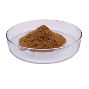 May include: A clear glass petri dish containing a mound of brown powder. The powder is a light brown color and appears to be finely ground. The dish is round and sits on a white background.