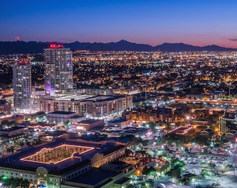 May include: An aerial view of a city at night with a mountain range in the background. The city is lit up with streetlights and buildings. There are several tall buildings in the foreground, including a hotel with a courtyard.