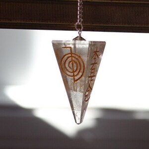 Puede incluir: Un péndulo de cristal de cuarzo transparente con una cadena de plata. El cristal tiene forma de pirámide y tiene un símbolo marrón grabado en un lado. El símbolo es una "H" estilizada con un círculo en el centro.