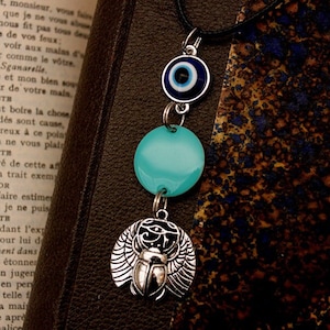 Peut inclure: Un collier avec trois pendentifs en argent : un œil bleu maléfique, un pendentif rond turquoise et un pendentif scarabée argenté avec des ailes.