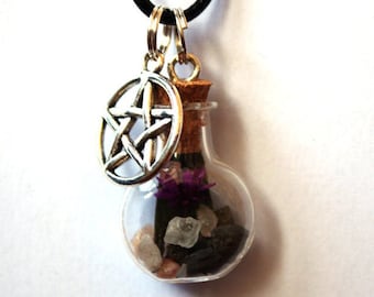 Puede incluir: Un collar con un pequeño colgante de botella de vidrio transparente relleno de piedras oscuras, cristales claros y una flor morada. La botella está sellada con un corcho y cuelga de un cordón negro junto a un dije de pentagrama de metal plateado.