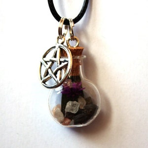May include: A necklace featuring a small clear glass bottle pendant filled with dark stones, light crystals, and a purple flower. The bottle is sealed with a cork and hangs from a black cord alongside a silver-toned metal pentagram charm.