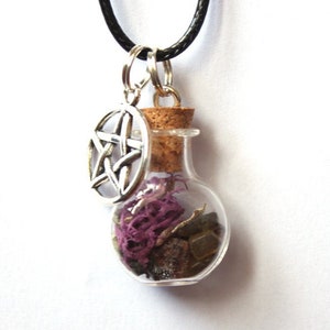 Puede incluir: Collar con colgante de vial de vidrio transparente con tapón de corcho, relleno de elementos morados y marrones. Un dije de pentagrama plateado cuelga junto al vial. El collar tiene un cordón negro.