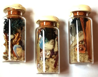 Puede incluir: Tres pequeños frascos de vidrio llenos de elementos naturales. Cada frasco tiene un tapón de corcho con una parte superior de color crema. El contenido incluye cristales, plantas secas y arena, creando una exhibición decorativa única.