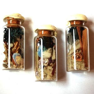 Könnte beinhalten: Drei kleine Glasflaschen gefüllt mit verschiedenen natürlichen Elementen. Jede Flasche hat einen Korken mit cremefarbenem Deckel. Der Inhalt umfasst Kristalle, getrocknete Pflanzen und Sand, wodurch eine einzigartige Dekoration entsteht.