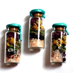 May include: Three small glass bottles with cork stoppers and teal wax seals. Each bottle is filled with dried flowers, herbs, and crystals. The bottles are arranged in a row on a white background.