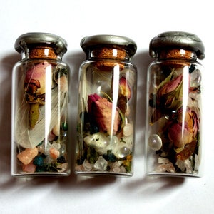May include: Three small glass bottles with cork stoppers and silver lids. Each bottle contains dried rose buds, crystals, and other decorative elements. The bottles are arranged in a row against a white background.