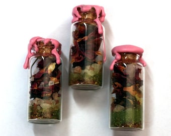 Puede incluir: Tres pequeños frascos de vidrio con tapones de corcho y sellos de cera rosa. Los frascos están llenos de flores secas, hierbas y cristales. El contenido es visible a través del vidrio transparente. Los frascos están colocados sobre un fondo blanco.