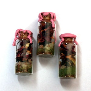 May include: Three small glass bottles with cork stoppers and pink wax seals. The bottles contain dried flowers, herbs, and crystals. The contents are visible through the clear glass. The bottles are set against a white background.