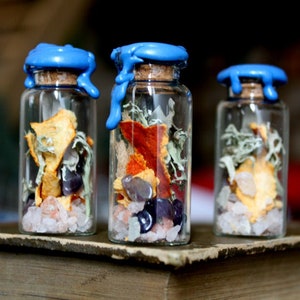 May include: Three small glass bottles with blue wax seals, filled with dried fruit, herbs, and crystals. The bottles are sitting on a wooden surface.