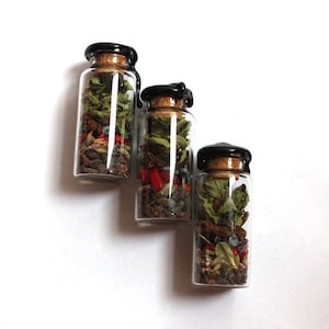 May include: Three small glass bottles filled with dried herbs and spices. Each bottle has a cork stopper sealed with black wax. The contents include green leaves, red peppers, and other colourful ingredients, creating a visually appealing arrangement.