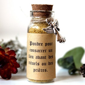 May include: A small glass bottle with a cork stopper and a silver angel charm. The bottle is filled with a light brown powder and has a brown paper label that reads "Poudre pour consacrer un lieu avant des rituels ou des prières."
