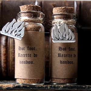 Peut inclure: Deux petites bouteilles en verre avec des bouchons en liège et des étiquettes en papier brun. Les étiquettes portent le texte "Hot foot. Recette de baudou." et un charme argenté en forme de flamme est attaché à chaque bouteille.