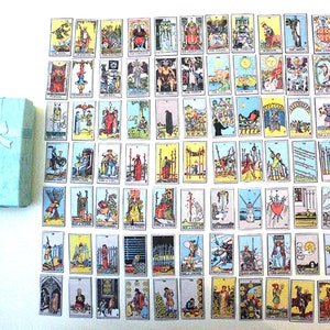 Peut inclure: Un jeu complet de cartes de tarot disposé sur une surface blanche. Les cartes sont disposées en grille et présentent des illustrations colorées. Une boîte cadeau bleu clair avec un ruban blanc se trouve dans le coin inférieur gauche de l'image.