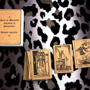 Può includere: Un set di carte dei tarocchi con cartoncino marrone e inchiostro nero. Le carte sono disposte su una superficie con stampa animalier bianco e nero. Le carte includono Il Matto, Il Mago e La Sacerdotessa. La scatola delle carte è marrone con il testo "Jarot de Marseille miniature et minimaliste. Arcanes majeurs."