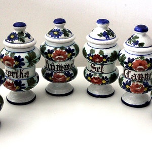 Può includere: Set di sei barattoli per spezie in ceramica bianca con disegni floreali blu e rossi. Ogni barattolo ha un'etichetta diversa: Paprika, Cumino, Sale, Carvi, Senape e Pepe.