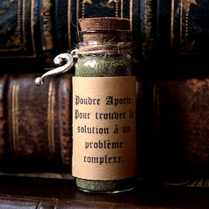 May include: A small glass bottle with a cork stopper and a brown paper label. The label reads "Poudre Aporie. Pour trouver la solution à un problème complexe." The bottle is filled with a green powder and has a silver question mark charm attached to the neck.