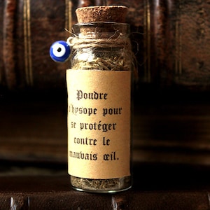Könnte beinhalten: Eine kleine Glasflasche gefüllt mit getrockneten Kräutern und einem Korkstopfen. Die Flasche hat ein Etikett mit der Aufschrift "Poudre d'hysope pour se protéger contre le mauvais oeil." Die Flasche ist mit einer Schnur gebunden und hat einen blauen Glücksbringer mit einem bösen Auge.