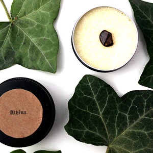 May include: A black tin with a brown label that reads "Athēna."  The tin is filled with a white substance and a brown stone is visible on top.  The tin is surrounded by green leaves.