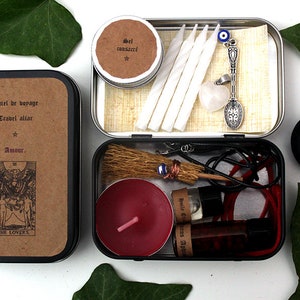 May include: A travel altar kit in a metal tin, featuring candles, a small broom, a spoon pendant, and a red candle. The tin includes text and a tarot card image. Additional items include a small container and a charcoal disc.